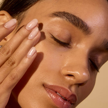 Close-up of a woman's face with one hand applying skincare, against a warm beige background.